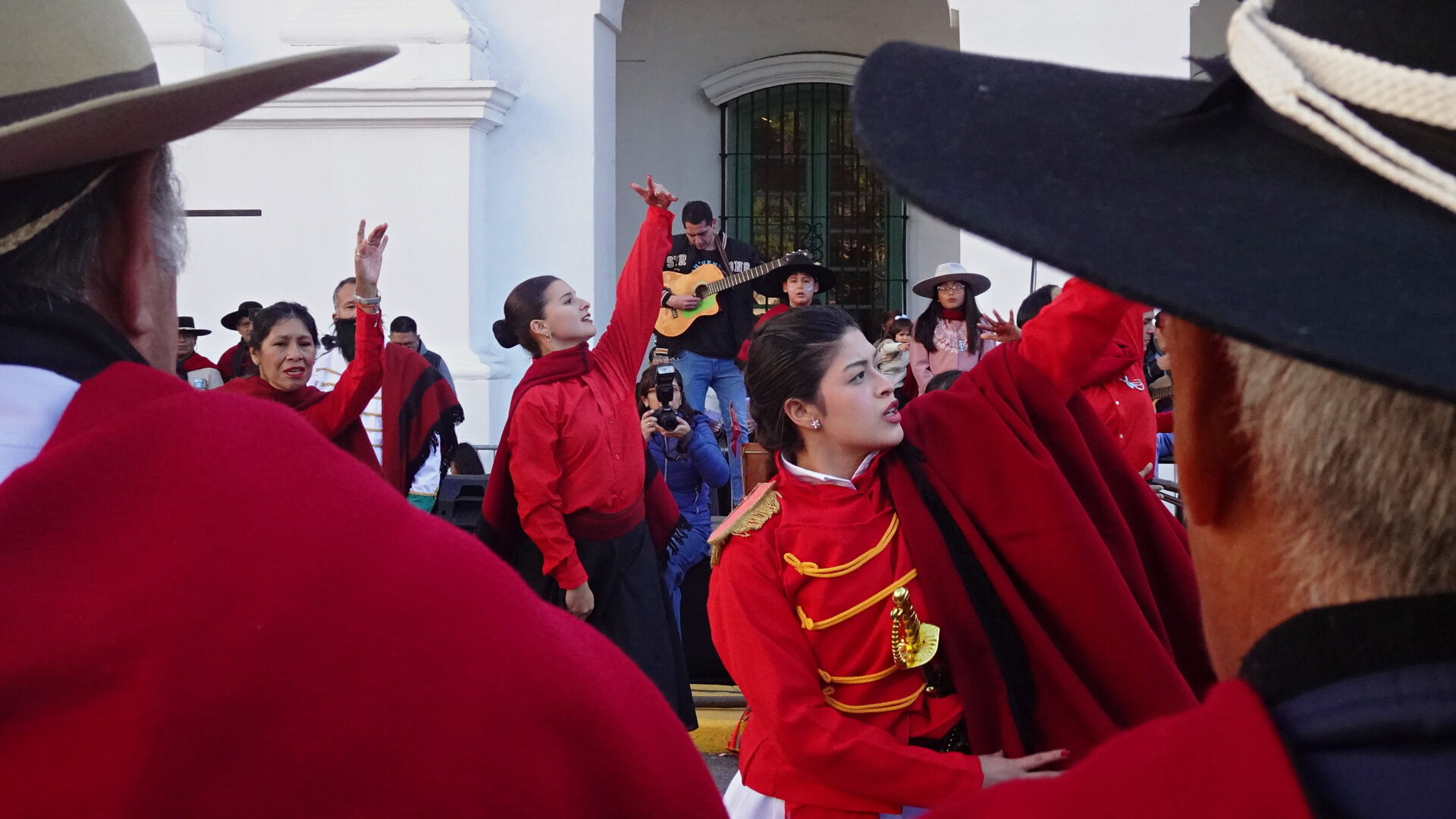 Dancers in red ponchos and wide-brimmed hats performing outside a white colonial building