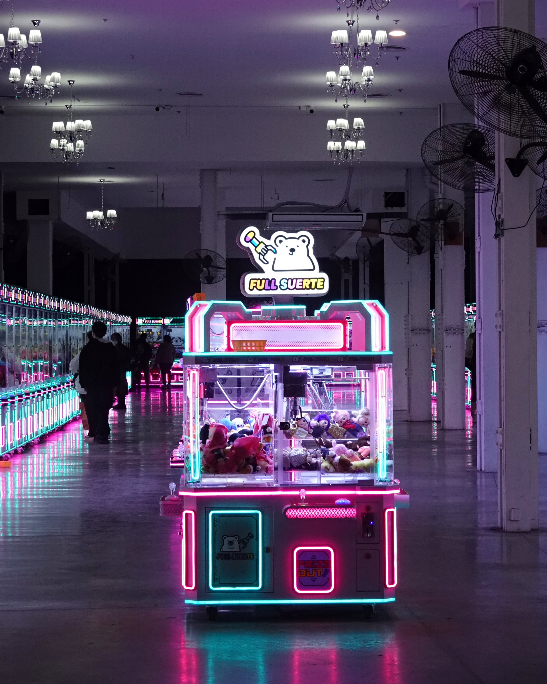 A neon-lit claw machine in a dim arcade hall with chandeliers