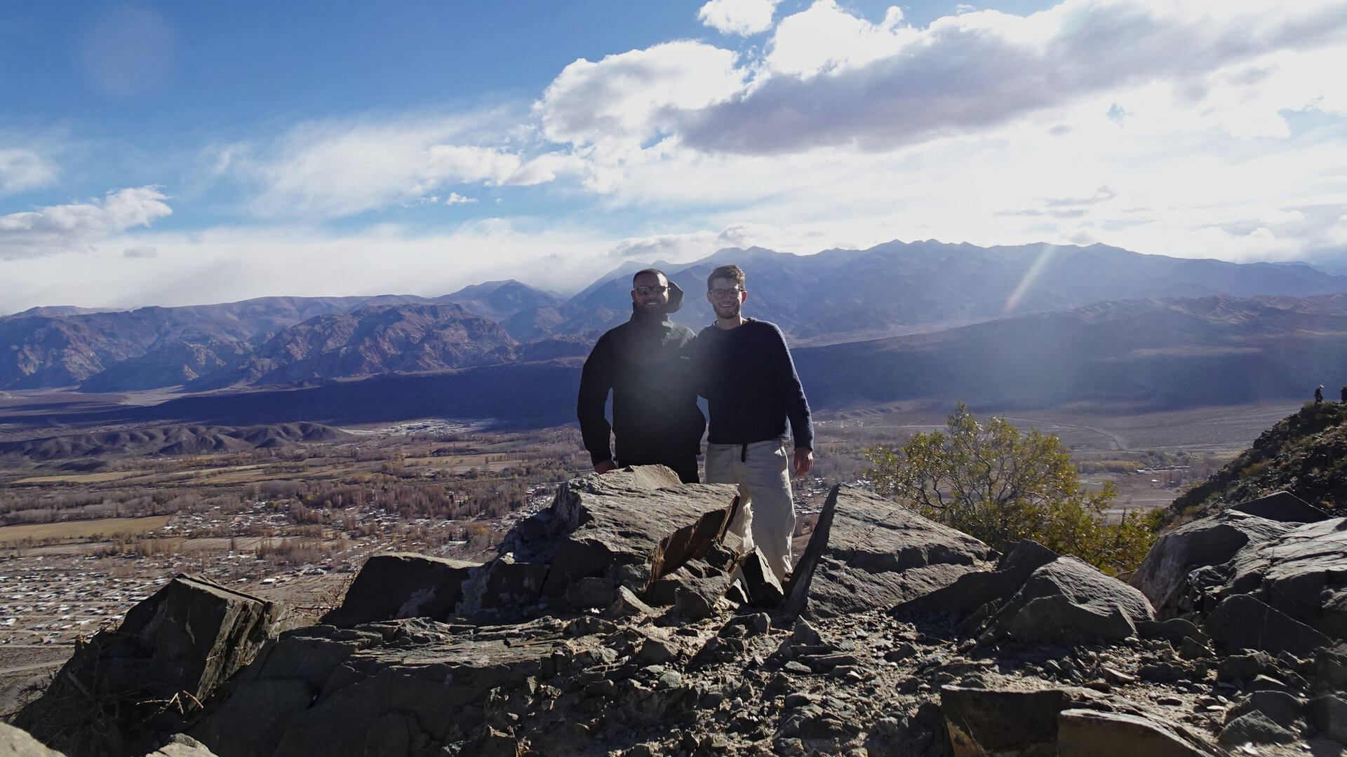 Two people on a rocky viewpoint above a valley town and distant mountains