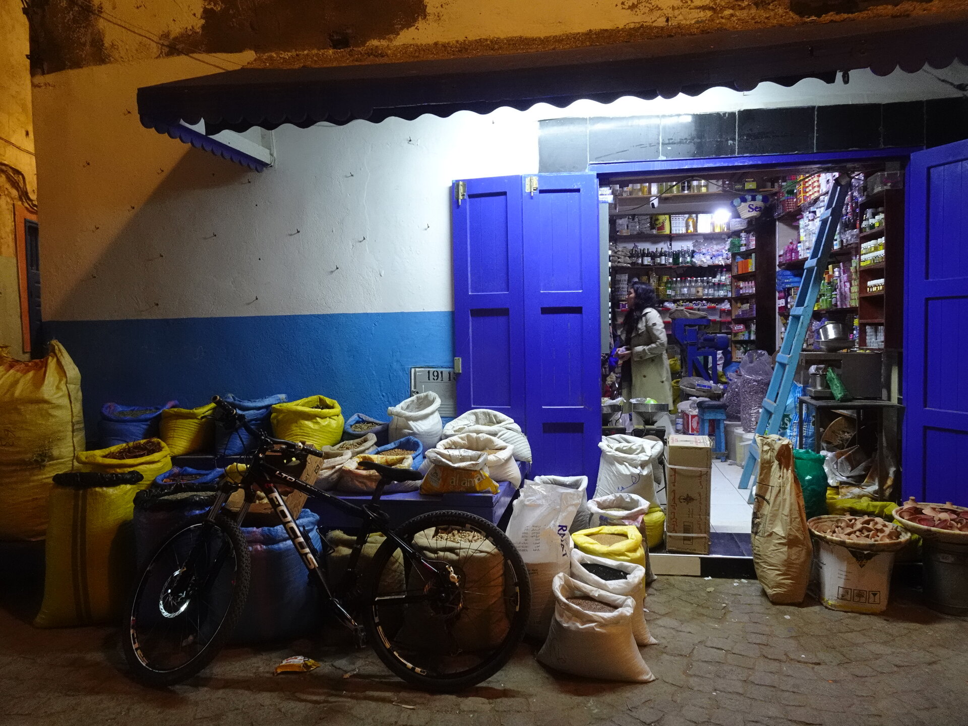 Bike parked among sacks of spices and grains outside a blue-doored shop at night
