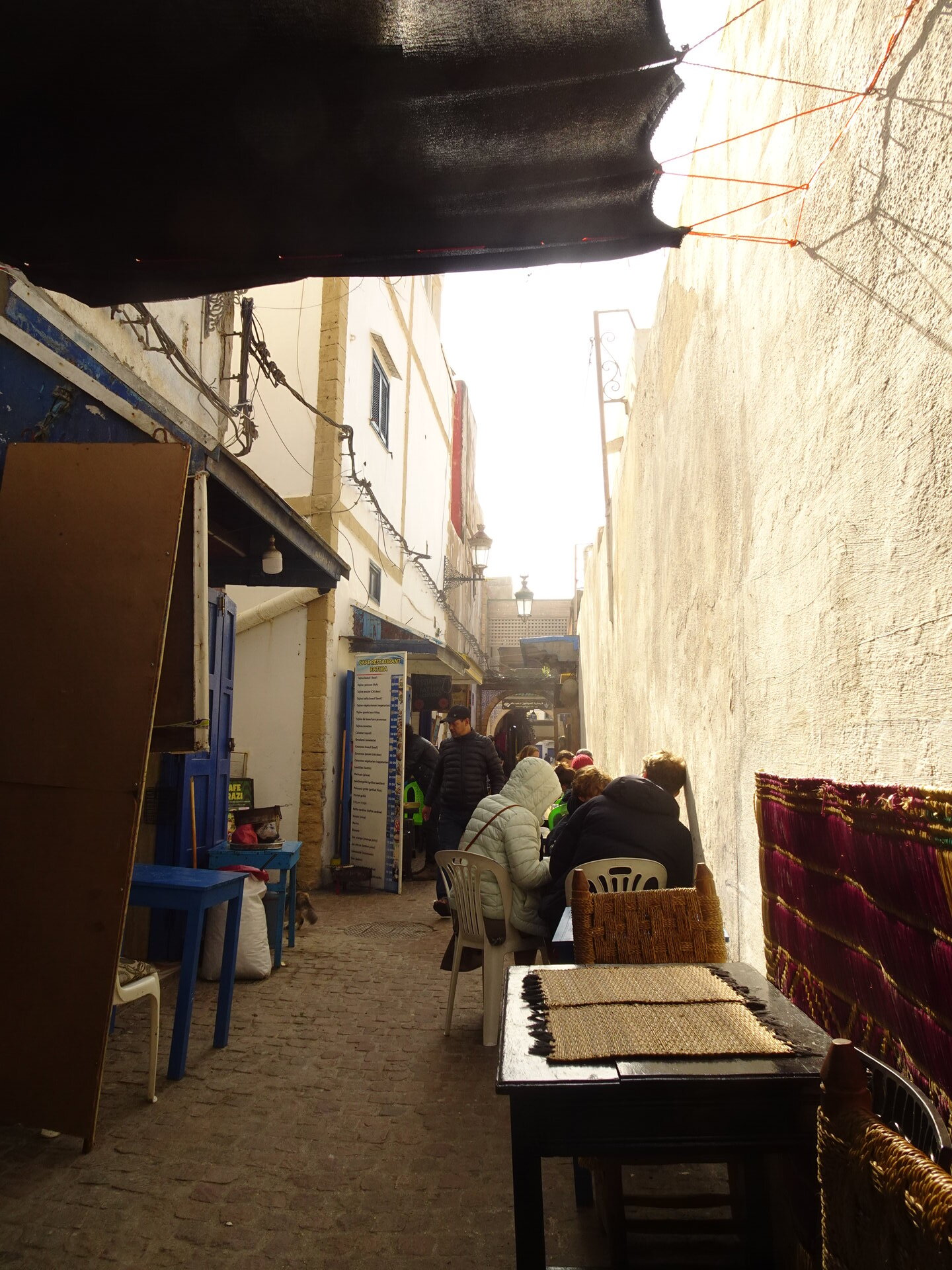 Shaded medina alleyway with cafe tables and people sitting in the afternoon sun