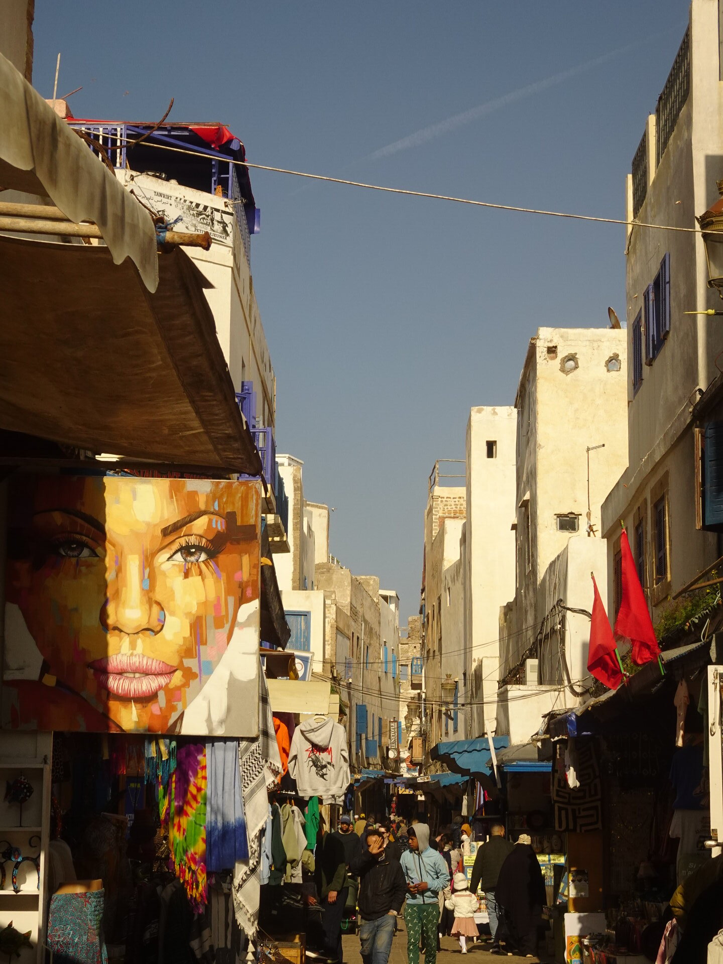 Busy medina street with a large face mural, market stalls, and Moroccan flags