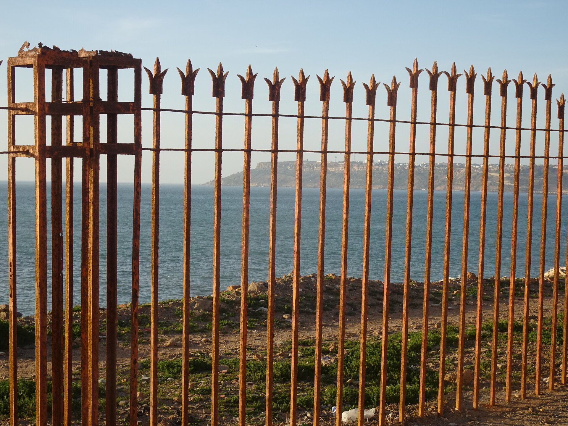Rusty iron fence with pointed tips overlooking the sea and a distant town