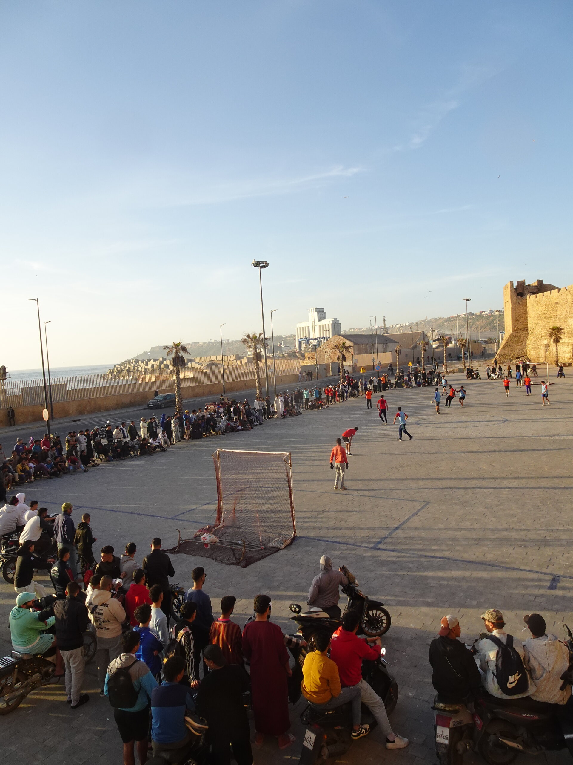 Crowd watching a street football match beside old city walls at sunset