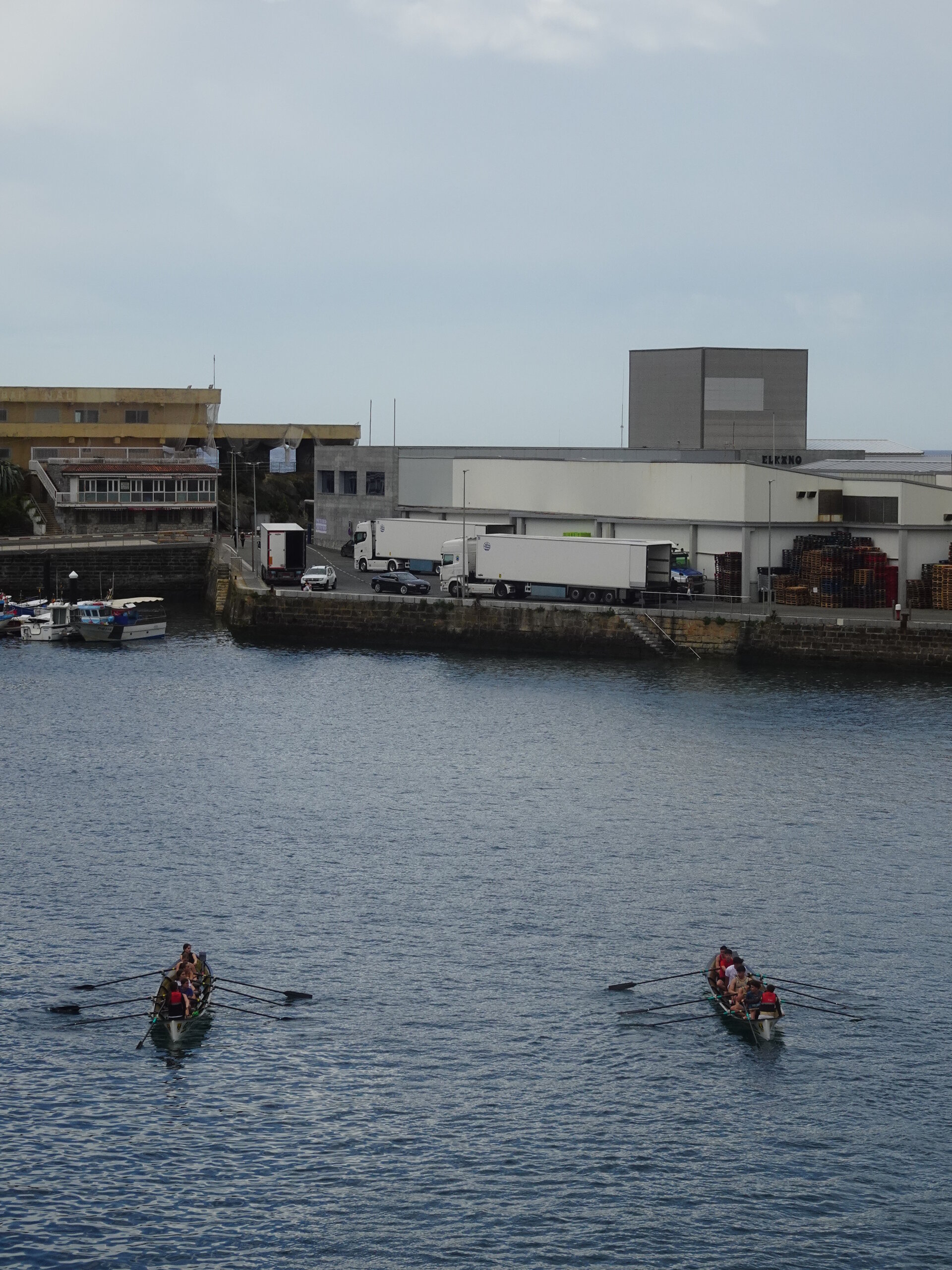 Two rowing crews on harbour water in front of dockside warehouses and trucks