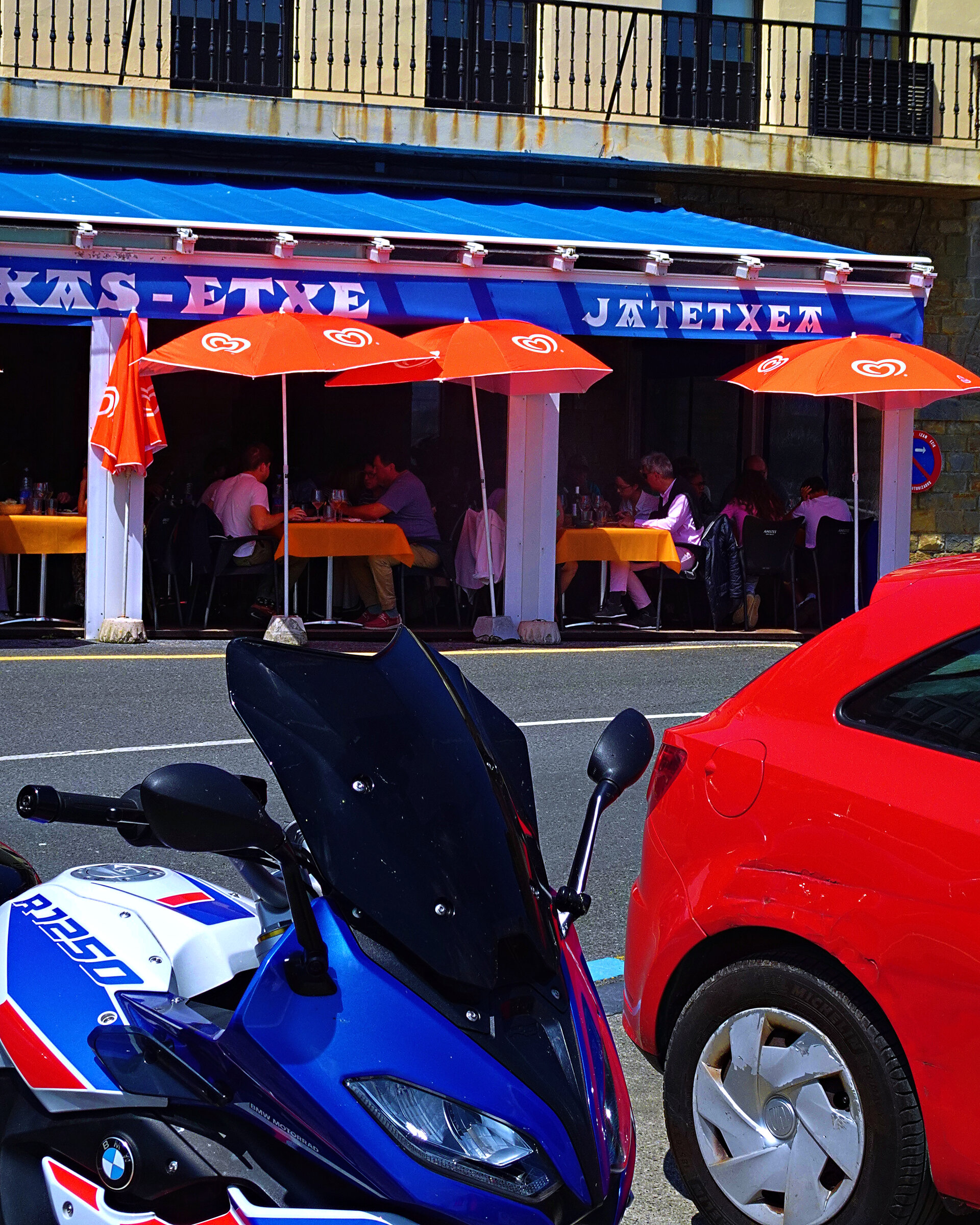 Orange parasols shading a busy harbourside restaurant terrace, motorbike and red car parked across the road