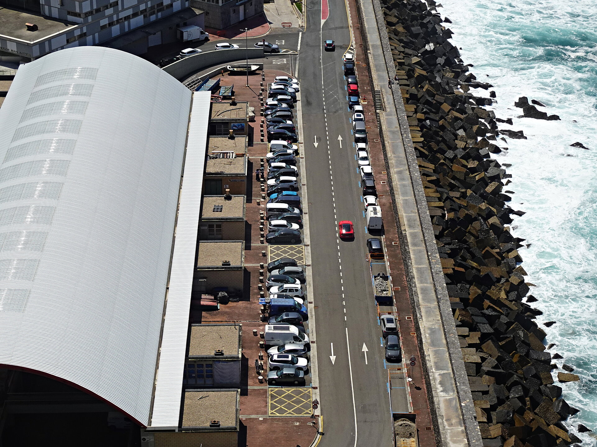 Aerial view of a seafront road and curved-roof building beside a rocky breakwater and surf
