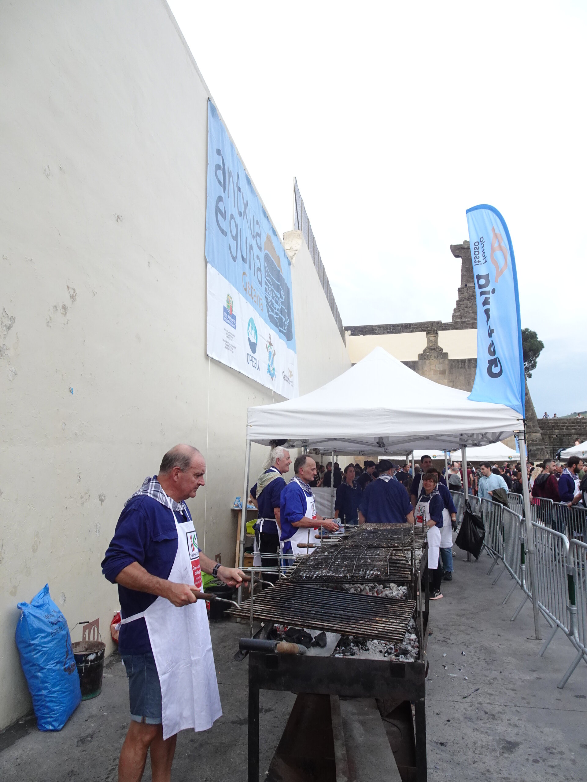 Men in aprons grilling over coals at a busy outdoor food festival