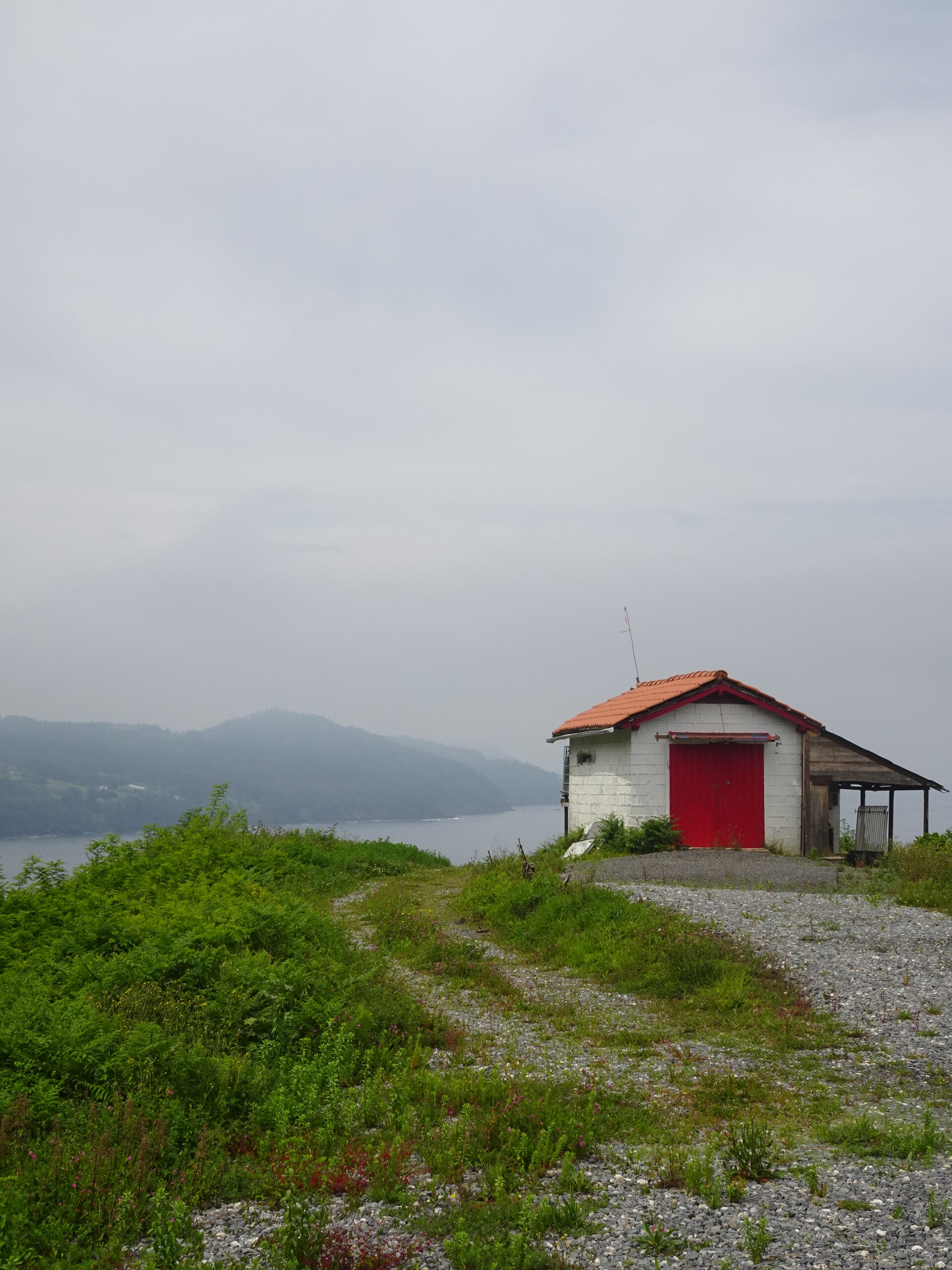 White boathouse with a red door on a headland above a misty bay
